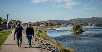 two people walking along a river walk with the river on the left and mountains in the background